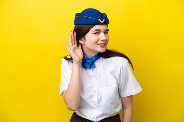 Airplane stewardess Russian woman isolated on yellow background listening to something by putting hand on the ear