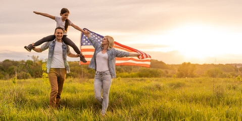 Young parents with their daughter holding American flag in countryside at sunset. Independence Day celebration