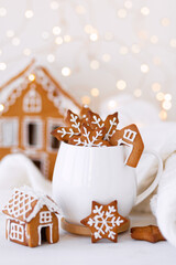 Hot winter drink in a white mug: cozy home composition with homemade gingerbread cookies, candy cane, fir tree branch. Wooden background, christmas lights and candles
