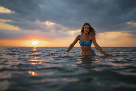 A girl with blond hair in a blue swimsuit splashes to the sides while sitting in an estuary on a sunset background