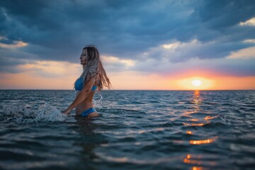 A girl with blond hair in a blue swimsuit splashes to the sides while sitting in an estuary on a sunset background