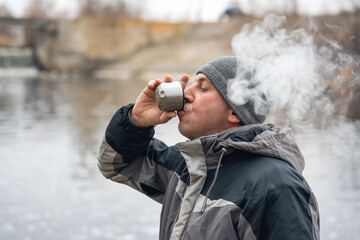 A man drinks hot coffee and smokes in nature, his face is covered with thick smoke - obrazy, fototapety, plakaty