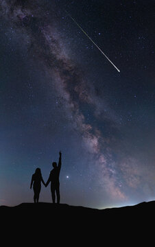 A couple of people in love hold hands and look at a shooting star at night against the backdrop of the Milky Way. The silhouettes are drawn by hand without a reference image. 