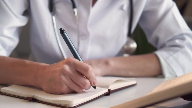 Medical education. Junior physician. Make notes. Unrecognizable woman writing information in notebook from encyclopedia sitting light room interior.