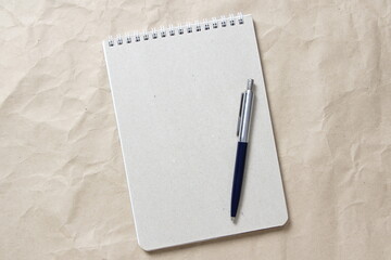 Gray notepad with white coiled spring and pen on a background of beige crumpled craft paper. With empty space for text and design