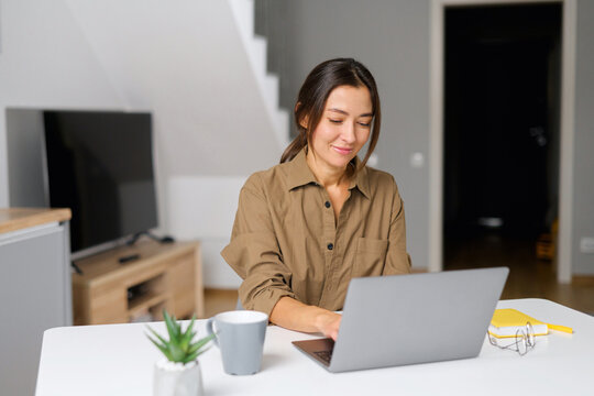 Smiling young joyful asian female freelancer enjoying morning coffee while working on the laptop, looking at the screen and sitting at the modern desk at home