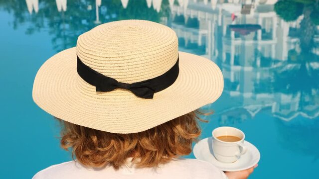 female in a straw hat holds a cup of coffee in her hand over a blue swimming pool concept of good morning and day planning, relaxation and enjoyment, back view