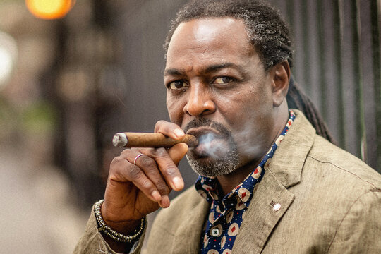 Fashionable closeup portrait of middle aged elegant afro American man wearing brown suit smoking cigar. Smiling and positive. Sitting on the stairs. Outdoor