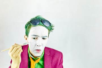 Mim is preparing for performance. Cosplayer guy paints his face with brush. Actor with white makeup and green hair in burgundy suit with yellow tie. Background with copy space for text.