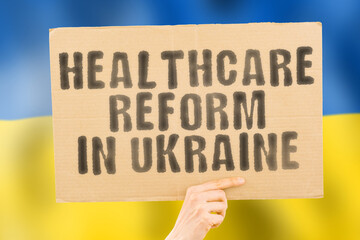 The phrase " Healthcare reform in Ukraine " on a banner in men's hand with blurred Ukrainian flag on the background. Nation. Crisis. Problem. Medical help. Reformation. Clinical