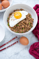 White bowl with fried rice and minced meat topped with a chicken egg, studio shot on a light-grey marble background