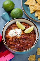 Chilli con carne or spicy meat stew topped with yogurt and lime wedges, vertical shot on a grey concrete surface, middle closeup