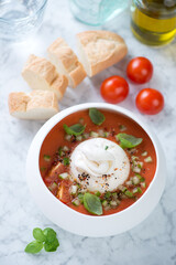 Bowl of tomato gazpacho soup with burrata cheese, green basil and baguette, elevated view on a light-grey marble background
