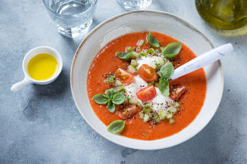 Plate of gazpacho made of red tomatoes and served with burrata and green basil, studio shot on a light-blue stone background