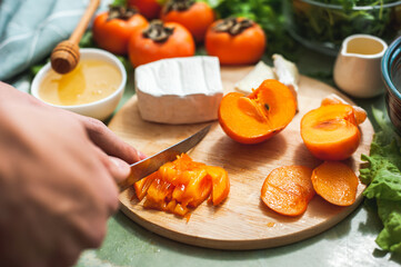 Ingredients and process of making a vitamin winter salad with persimmons, tangerines and cheese Female or teenage hand cuts persimmon on a round wooden board Vegan lunch preparation. Selective focus