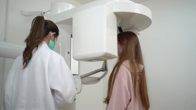 A cute long-haired Caucasian teenage girl walking into a digital dental x-ray machine and rest teeth in the bite tab, getting a 3D panoramic teeth x-ray controlled by Asian female dentist in face mask