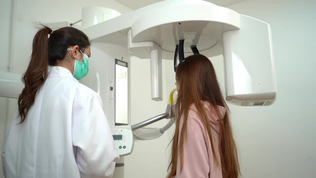 Cute long-haired Caucasian teenage girl standing taking a 3D panoramic teeth x-ray with a digital dental x-ray machine, then watching a teeth x-ray on tablet shown by Asian female dentist in face mask