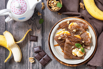 Celebrating Pancake day, healthy breakfast. Delicious homemade chocolate crepes with nuts, caramel, bananas on rustic wooden table. Top view flat lay.