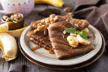 Celebrating Pancake day, healthy breakfast. Delicious homemade chocolate crepes with nuts, caramel, bananas on rustic wooden table.