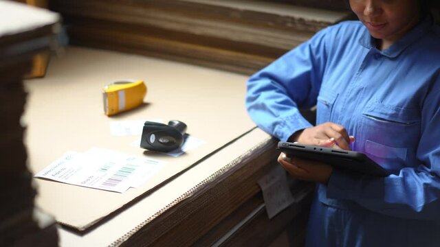 Cropped video of female warehouse worker in blue uniform using wireless barcode scanner scanning barcode or label on paperwork placed on flat cardboard sheet and record the data on tablet in warehouse