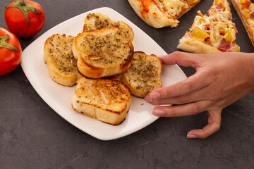 hand taking a crispy garlic bread with oregano, in the background slices of pizza with cheese, pineapple and ham, decoration with tomatoes, table reserved and ready to eat with appetizer, restaurant a