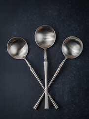 Three crossed brushed stainless steel soup spoons on a dark stone background. Close-up.