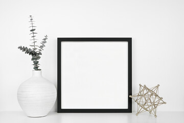Mock up black square frame with modern home decor. White shelf against a white wall.  Eucalyptus...