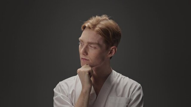Portrait Of Young Guy In Medical Gown. He Has Red Hair And Freckles. Guy Touches His Chin With His Hand. He Looks Up With Pensive Look.