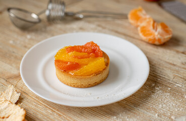 A tartlet with tangerine and grapefruit lies on a plate on a wooden table, around the tools and ingredients for making a dessert