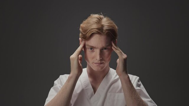 Portrait Of Young Guy In Medical Gown. He Has Red Hair And Freckles. Guy Massages His Temples With His Hands.