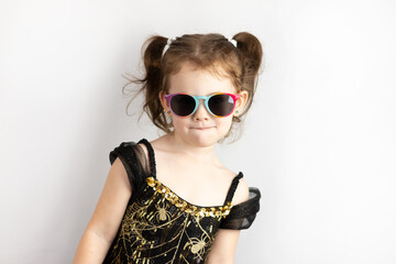 Naklejka premium Little cheerful girl with two ponytails and a black and gold dress in sunglasses. Studio photo on white of a grimacing child