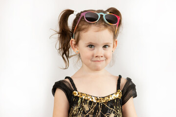 Naklejka premium Little beautiful girl with two ponytails and a black and gold dress. Studio photo on a white background of a child with sunglasses on his head
