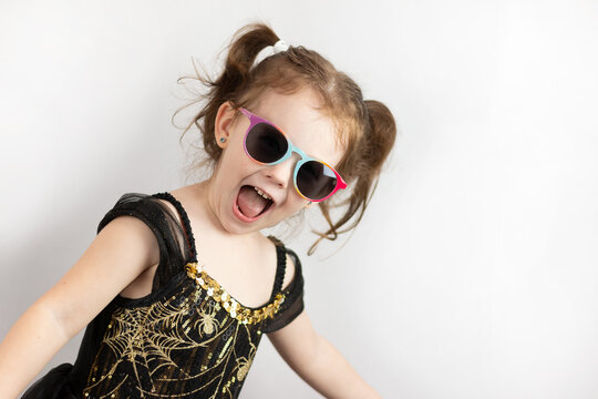 Little cheerful girl with two ponytails and a black and gold dress in sunglasses. Studio photo on white of a fooling child