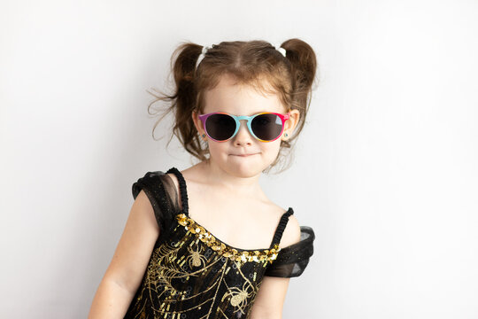 Little cheerful girl with two ponytails and a black and gold dress in sunglasses. Studio photo on white of a grimacing child