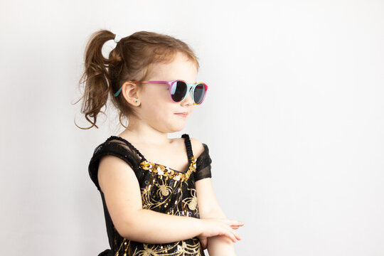 Little cheerful girl with two ponytails and a black and gold dress in sunglasses. Studio photo of a cute baby on a white background