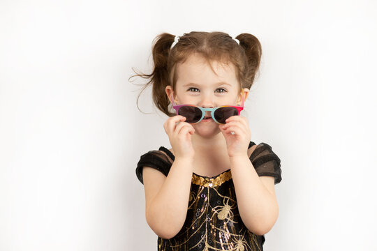 A little beautiful girl with two ponytails and a black and gold dress takes off her sunglasses smiling. Studio photo isolated on white background