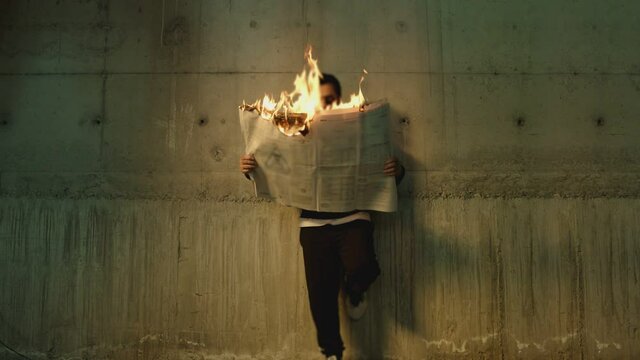 Footage of male artist with sunglasses standing near wall , holding and reading newspaper during it burns . Concept or symbol of fake or forbidden news   . Man reads burning magazine . Slow motion