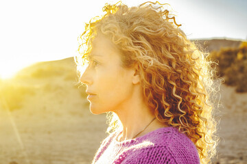 Sunset woman portrait looking serious. Curly blonde long hair female people enjoy golden sun light. Adult lady face expression.
