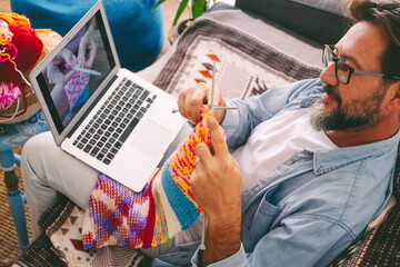 Handsome adult man learning how to knitting work with woolen yarn at home watching an online class...