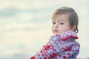A toddler sits on a stone beach. A cute baby girl on a beautiful sweater on the shore of the winter...