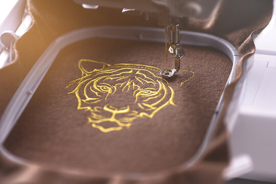 modern embroidery machine stiches a luxurious tiger head on brown merino wool fabric with golden yarn in sunny surrounding. fashion and chinese new year celebration concept. selective focus