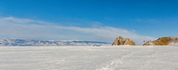 View of the frozen Lake Baikal and the mountains on a winter sunny day. Sacred Shamanka Mountain on Olkhon Island. Tourists walk on the ice, admire the beauty of the ice. Panorama.