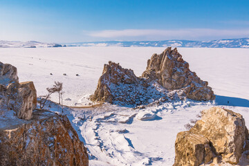 Sacred Shamanka Mountain on Olkhon Island in winter. View of the frozen Lake Baikal on a sunny day. View from above. Below, tourists walk on the ice - 480615980