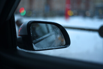Snow covered car rear view mirror on bokeh winter cityscape background.