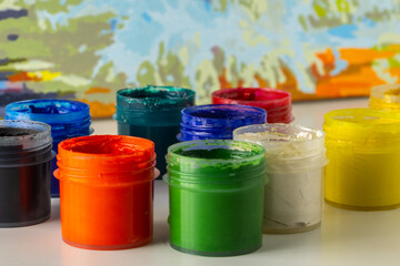 Open jars of multi-colored gouache on a blurred watercolor background. The concept of learning to draw. Materials for creativity.