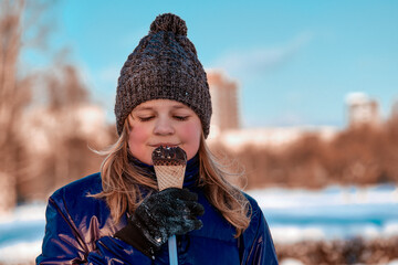 The child eats ice cream outdoors in the park in winter. A cute ten-year-old girl is holding a waffle cup with ice cream , covered in chocolate.