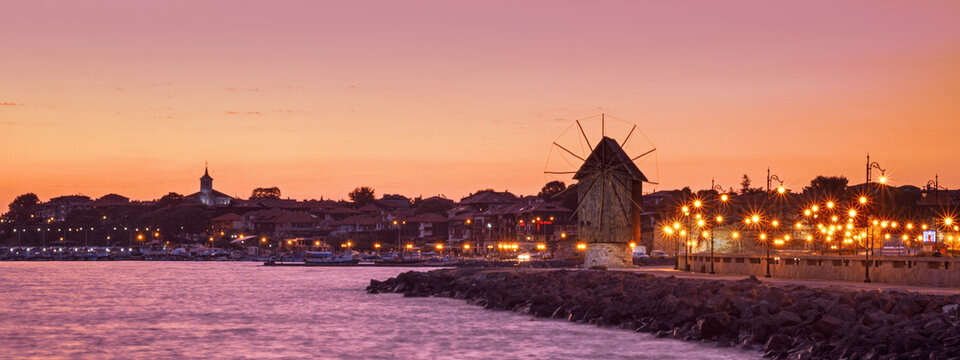 Evening city landscape, banner - view of the embankment with street lights and the wooden windmill before the entering to the Old Town of Nessebar, on the Black Sea coast of Bulgaria