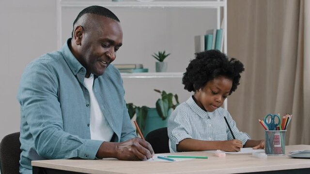 Mature african american father with little kid girl sitting in room at desk draws colored pencils dad and daughter play compete for best drawing studying doing homework happy fatherhood home hobby
