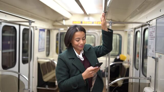 Businesswoman in subway carriage using smartphone to send business email or text with family. Young african american female travel by public transport standing in empty underground wagon late at night