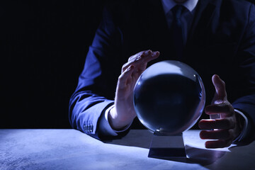 Businessman using crystal ball to predict future at table in darkness, closeup. Space for text